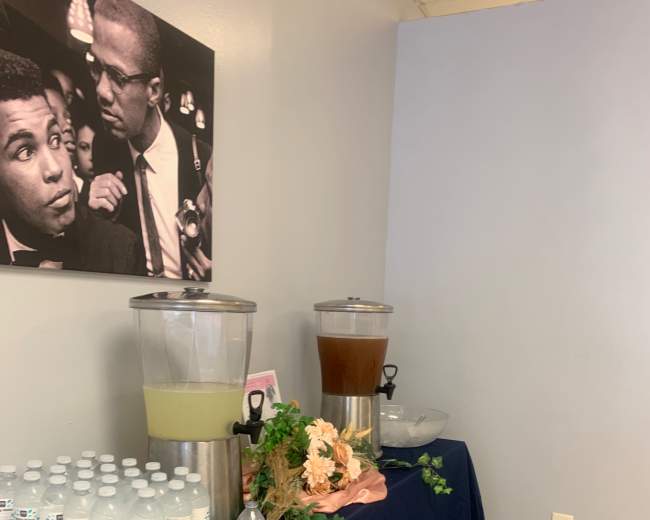 A table with two beverage dispensers, one containing a light yellow drink and the other a dark liquid, alongside a stack of bottled water and a floral arrangement.