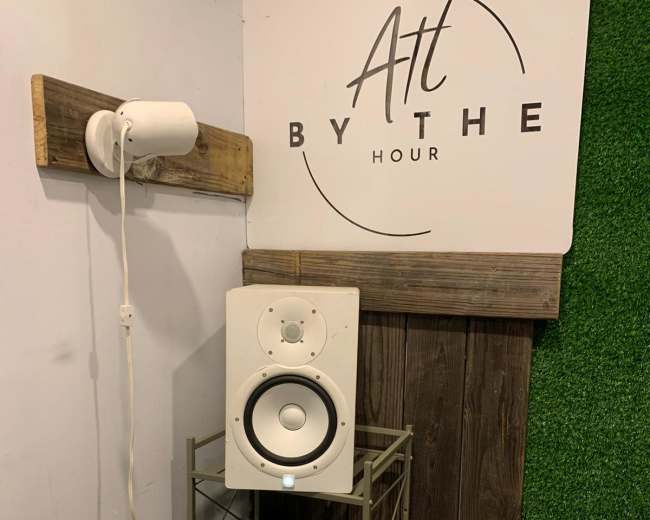 A white speaker is placed on a metal shelving unit next to a sign that reads "Art By The Hour," against a wooden and green backdrop.