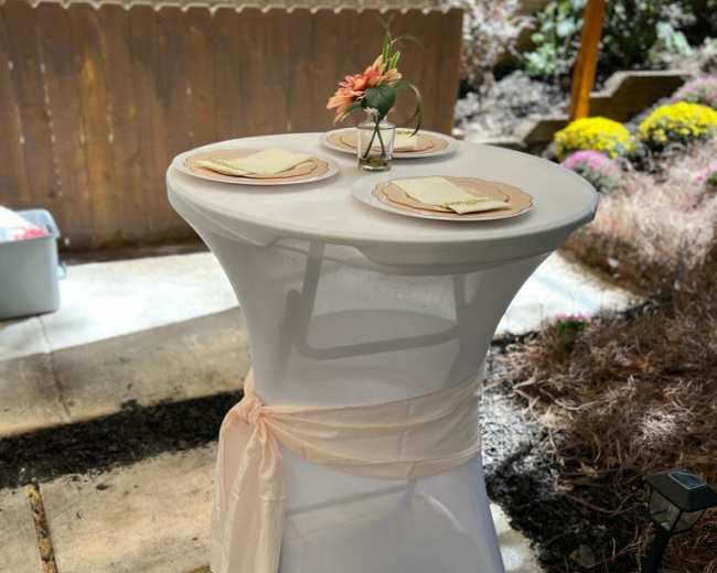 A small round table dressed in a white tablecloth is set with two plates and a flower vase in an outdoor garden setting.