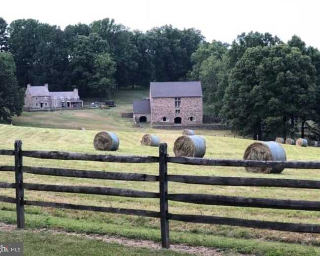 A rural landscape with a stone farmhouse and barn in the background, surrounded by round hay bales and a wooden fence.