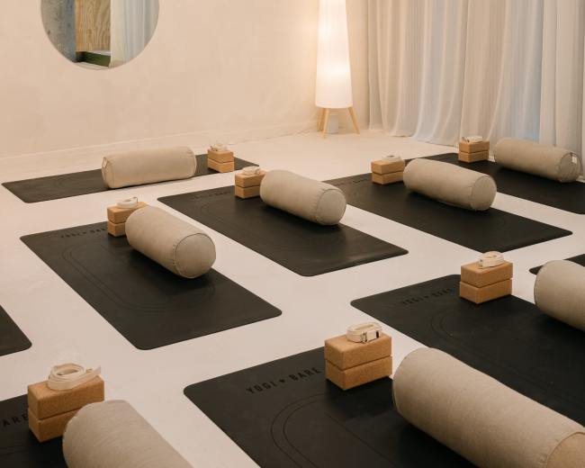 The image shows a minimalist yoga studio with rolled mats arranged on the floor, a tall lamp in the corner, and a circular mirror on the wall.