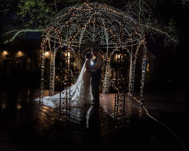 A couple stands under a lit gazebo at night, surrounded by a reflective wooden deck and trees.
