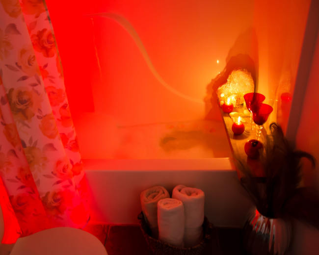 The bathroom features a bathtub illuminated by red lighting, surrounded by decorative towels and candles on a wooden shelf.