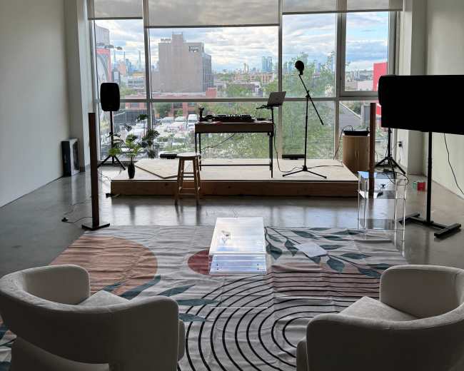 The image shows a spacious interior with two white chairs, a wooden platform with musical equipment, and large windows offering a view of a city skyline.