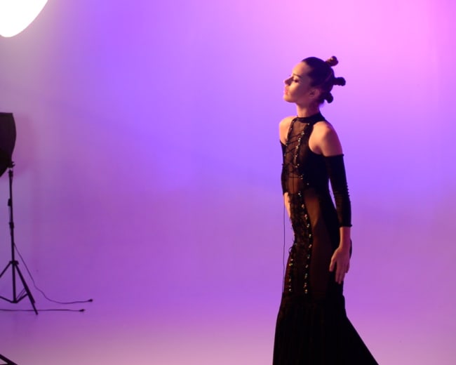 A model poses under a large studio light while wearing a black evening gown against a purple background.