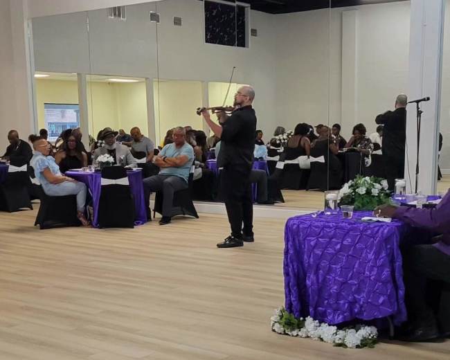 A man plays the violin in a banquet hall filled with seated guests at elegantly set tables.