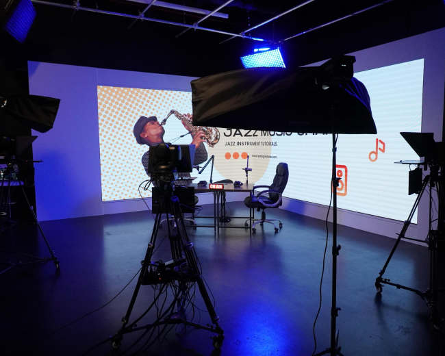 The image shows a television studio setup with multiple cameras facing an empty discussion area featuring a large screen displaying "JAZZ MUSIC NIGHT."