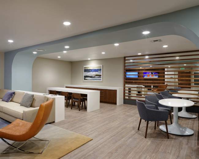 The image shows a modern lounge area with a sofa, chairs, and tables, featuring a light-colored wooden floor and subtle lighting.