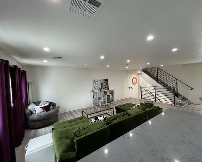 The image shows a modern living room with a green sectional sofa, a gray armchair, and a staircase with a metal railing in the background.