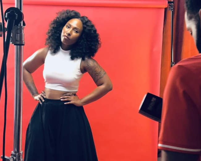 A woman poses with her hands on her hips in front of a red background during a photoshoot.