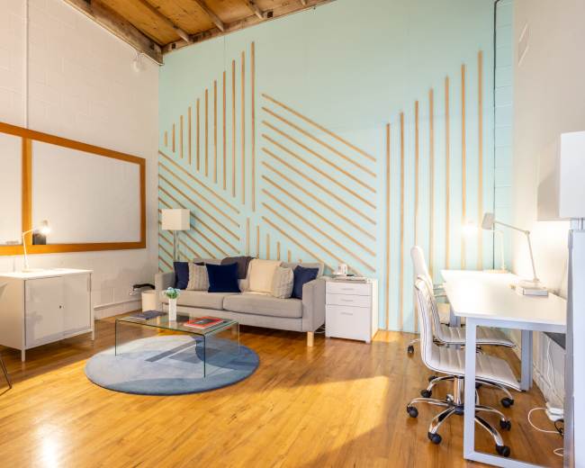 A modern workspace featuring a gray sofa, white desks with chairs, a round rug, and a textured wall accentuated with wooden stripes.