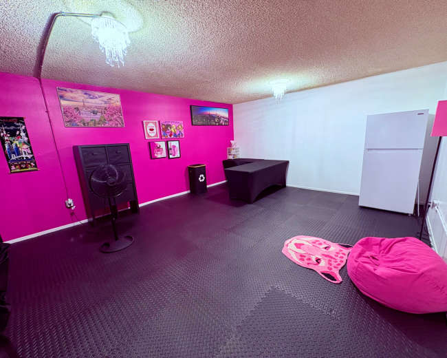 The image shows a room with pink walls, black rubber flooring, a table, a refrigerator, and a pink bean bag chair.