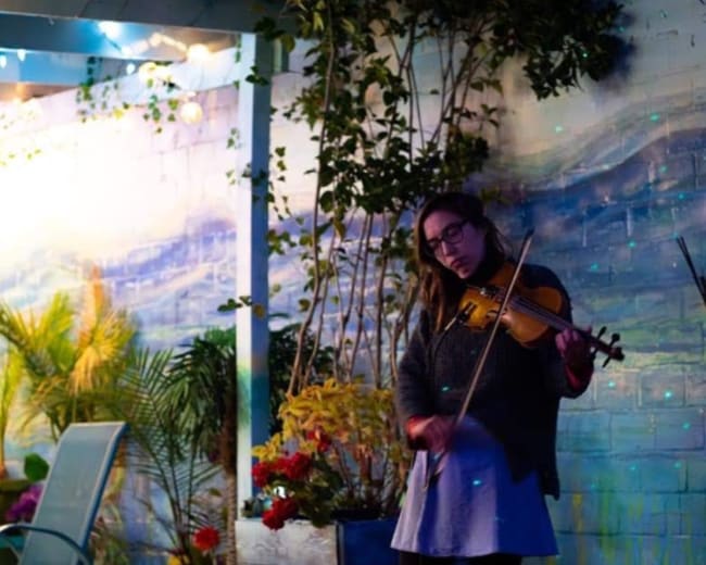 A woman plays a violin in front of a painted mural surrounded by plants and colorful lighting.