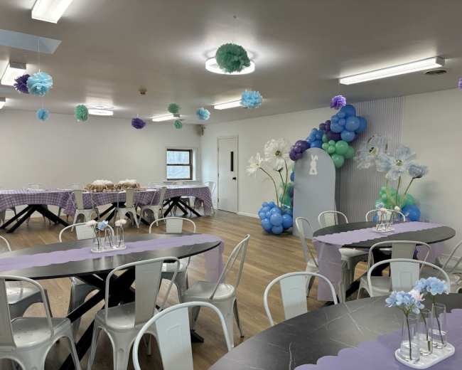 The image shows a decorated room with tables set up for an event, featuring pastel balloons and paper decorations hanging from the ceiling.