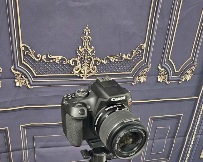 A Canon camera is mounted on a black tripod in front of a patterned backdrop.