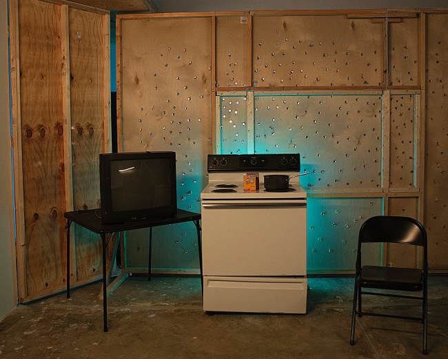 A simple kitchen setup features a stove, a small table with a television, a chair, and a plywood wall with drilled holes in a dimly lit room.