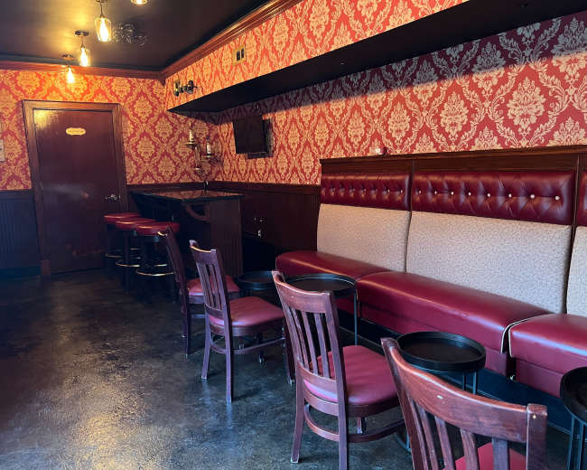 The image shows a sparsely furnished bar area with red wallpaper, a booth along one wall, and wooden chairs around small tables.