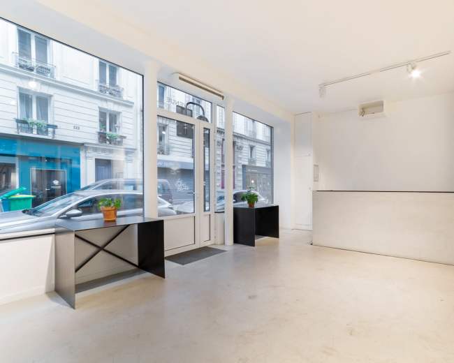 The image shows a minimalistic interior of a storefront with large windows, a reception counter, and two black pedestals.