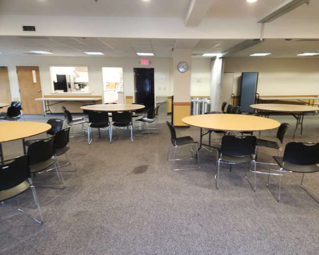 The image shows a large vacant room furnished with several round tables and chairs, with a kitchenette visible in the background.