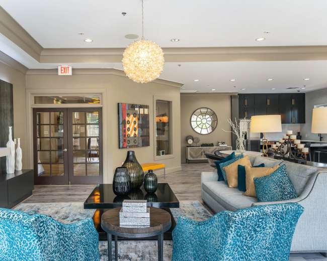 The image shows a modern, stylish lounge area featuring a seating arrangement with teal-patterned chairs, a central coffee table, decorative vases, and a large circular window.