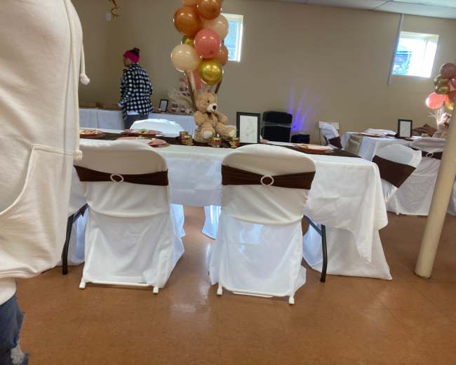 A decorated table with white tablecloths and brown sash chairs is set up in a room, featuring a teddy bear and balloons as centerpieces.