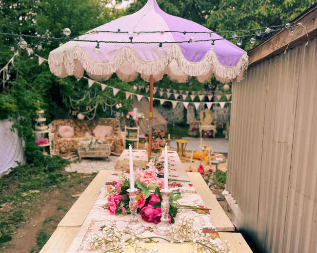 A long outdoor table is set with floral arrangements and candles under a decorative purple umbrella, surrounded by a whimsical garden setting.