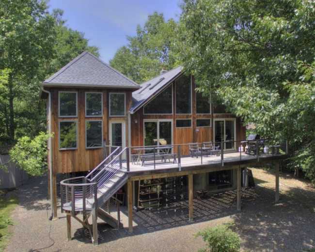 A wooden house with large windows and a wraparound deck is situated among trees.
