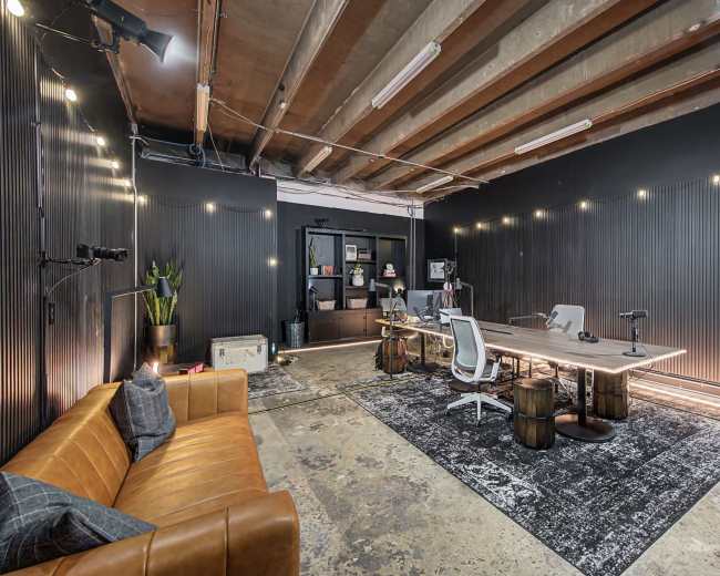 The image shows a modern office space with a large wooden table, several white chairs, a brown leather sofa, and ambient lighting against black walls.