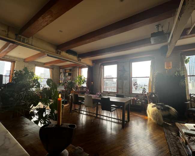 A spacious room features wooden beams, large windows, and a mix of dining and lounging areas, surrounded by plants and natural light.