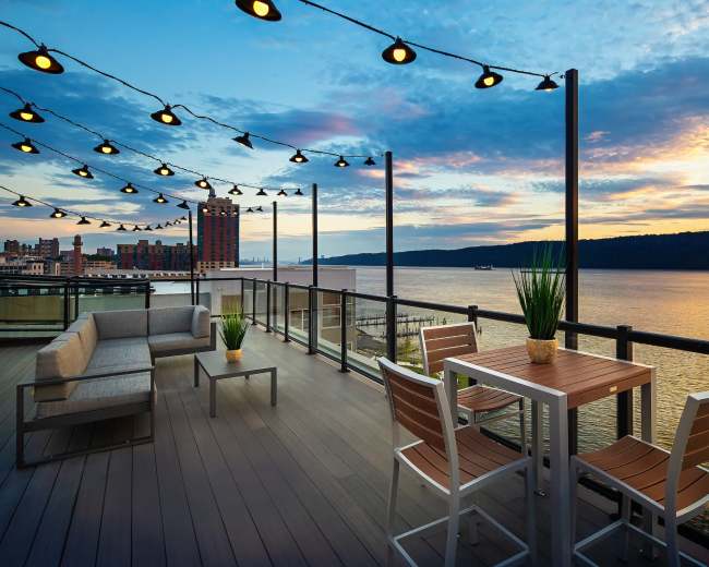 A modern balcony overlooks a river at sunset, adorned with string lights and stylish outdoor furniture.