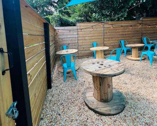 The outdoor space features several wooden tables and blue chairs surrounded by wooden fencing and shaded by a blue tarp.