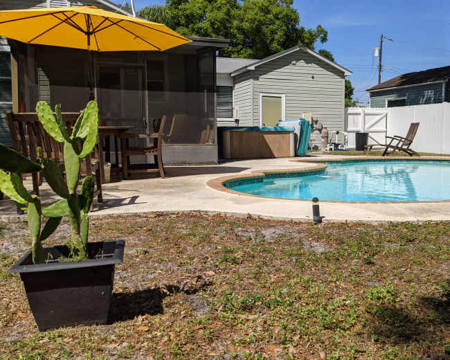A swimming pool surrounded by a patio, outdoor furniture, and a yellow umbrella, with a house and landscaped area in the background.