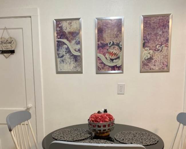 A round table with two chairs is set against a white wall adorned with three framed artworks, with a bowl of flowers in the center of the table.