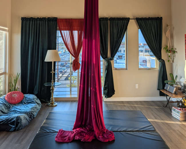 The image shows a performance space featuring a burgundy aerial silk suspended from the ceiling, with large windows allowing natural light and several curtains in various colors.