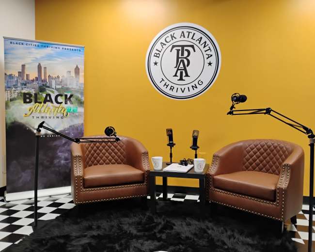 The image shows a cozy podcast studio with two brown leather chairs, two microphone stands, and a backdrop featuring a yellow wall with the logo "BLACK ATLANTA Thriving."