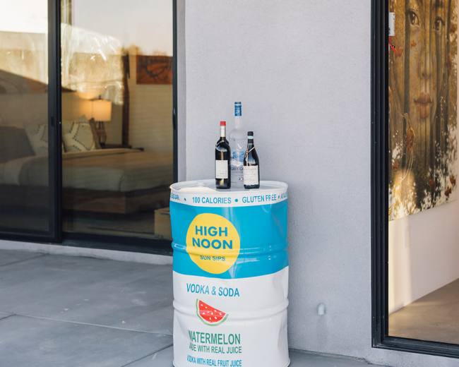 A colorful beverage cooler stands outside a modern building, featuring bottles of vodka and soda alongside a container marked for watermelon-flavored drinks.