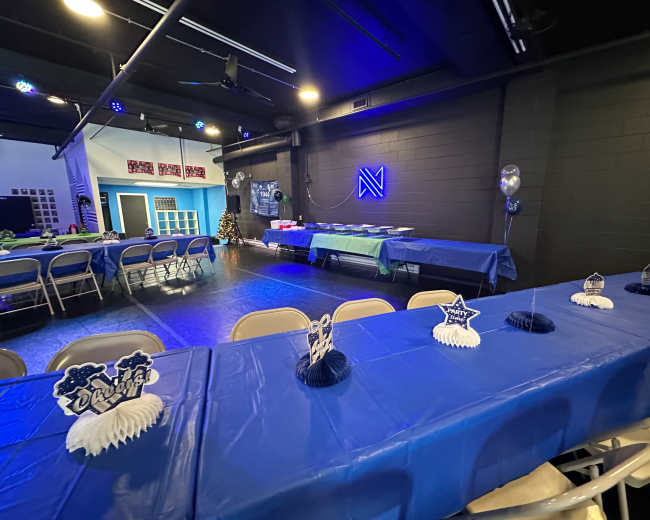 The image shows a decorated event space with blue tables set up for a gathering, featuring centerpieces and a buffet table in the background.