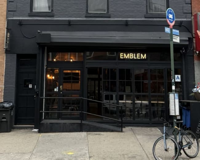 The image shows a three-story gray building featuring large windows and a storefront with the name "EMBLEM" illuminated above the entrance.