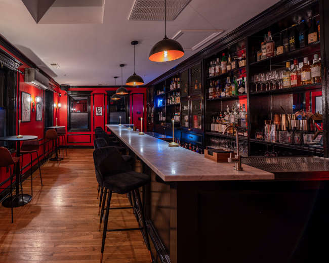 The image shows a dimly lit bar with red walls, a marble-topped counter, and shelves stocked with various bottles of liquor.