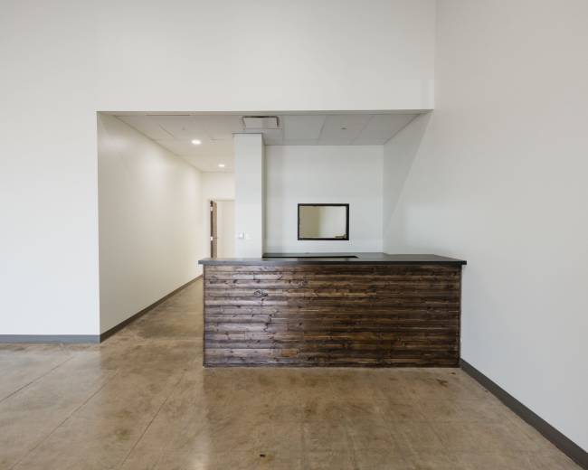 The image shows a minimalist reception area with a wooden counter and a mirror on the wall behind it, leading to a plain hallway.