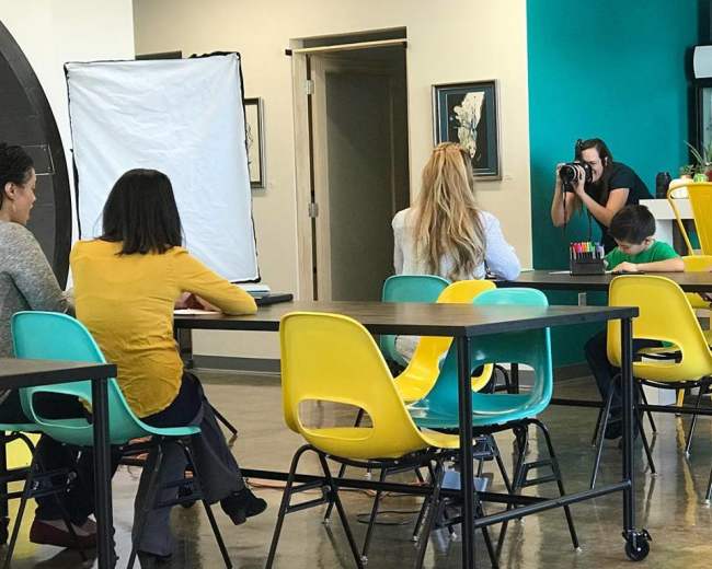 A group of individuals is seated at a table in an indoor space, with one person positioned in front of a camera being photographed.