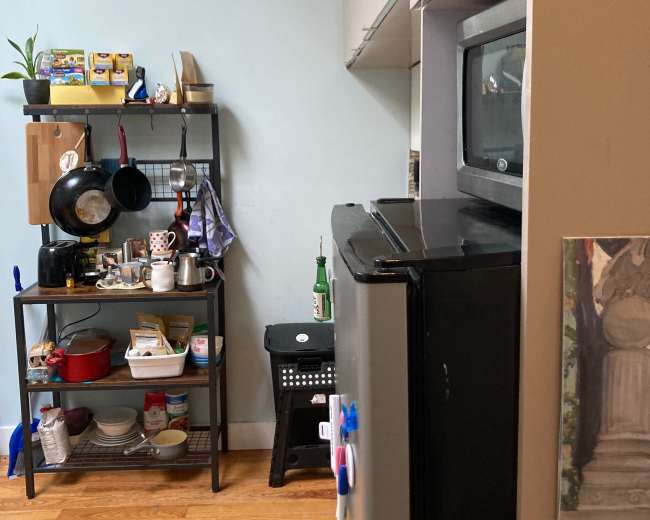 The image shows a kitchen area with cabinets, a black refrigerator, a microwave, and a storage cart with various kitchen items.