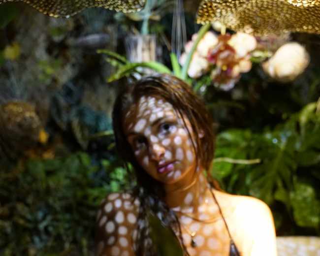 A woman sits in a bath surrounded by plants, with intricate shadows cast across her face and body from overhead lighting.