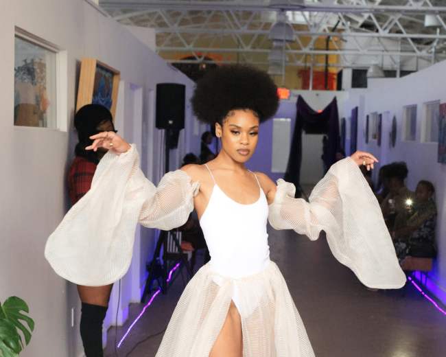 A model walks down a narrow hallway wearing a white bodysuit and a flowing, sheer skirt.