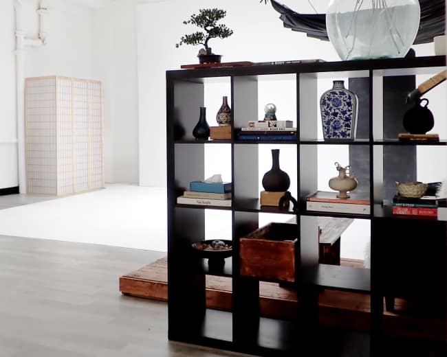 The image shows a spacious, minimalist interior with a black bookshelf containing decorative items and a large vase with flowers, while natural light enters through a skylight.