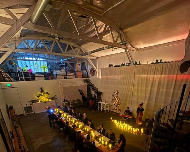 A long table set with candles occupies a spacious, dimly lit event space featuring a staircase and decorative elements in the background.