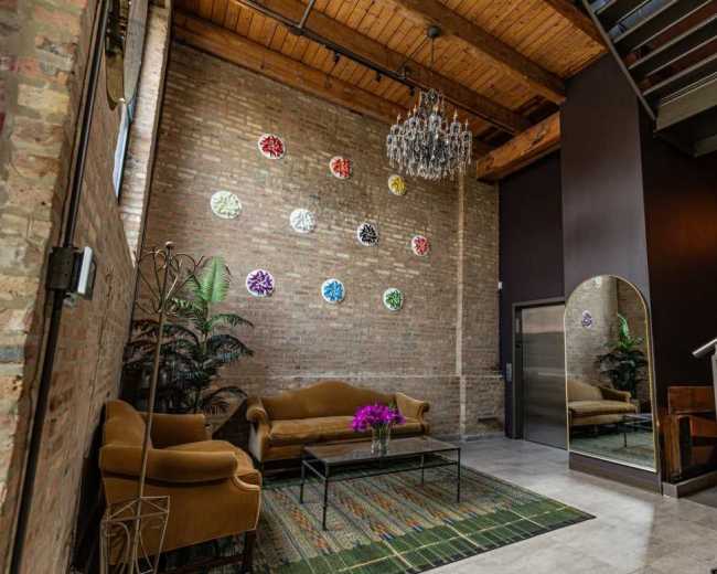 A cozy entryway features a brown sofa, a coffee table with flowers, a mirror, and colorful wall decor against a brick wall.