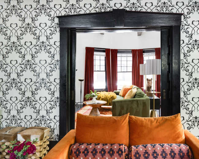 The image shows a cozy living room viewed through an ornate doorway, featuring patterned wallpaper, vibrant furniture, and large windows with red curtains.