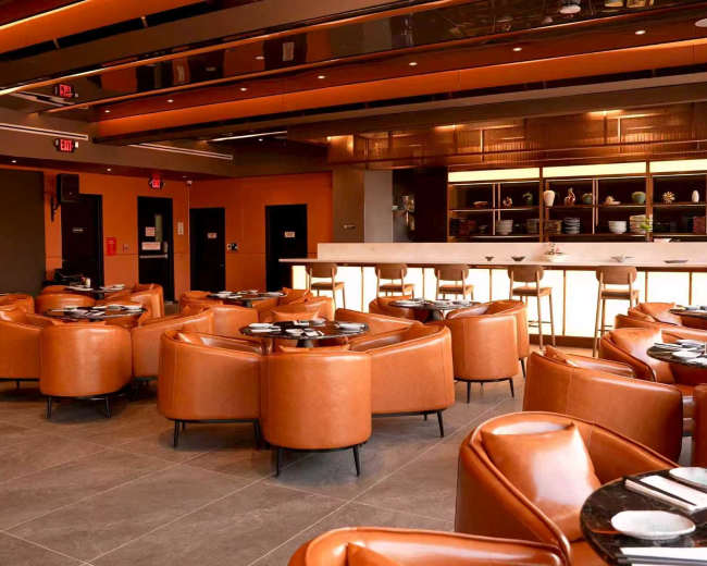 The image shows a modern restaurant interior featuring brown leather seating, a central bar area, and a sleek minimalist design.