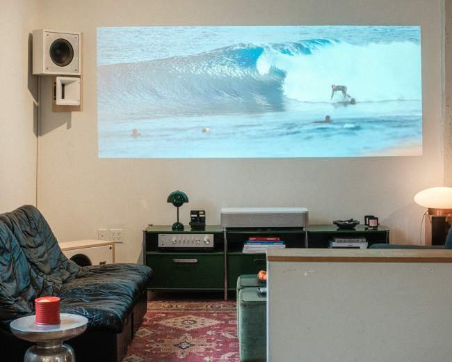 A living room features a projection of a surfer riding a wave on one wall, with a sofa, coffee table, and green storage units nearby.